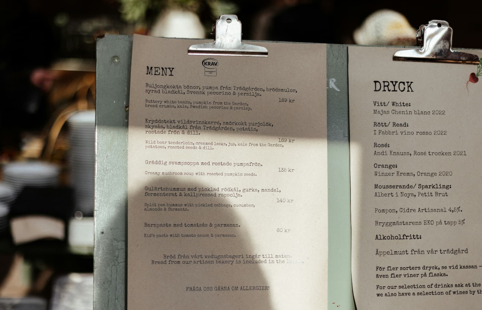 Printed food and drinks menus clipped to a display board inside a restaurant, with dishes listed in Swedish and English.