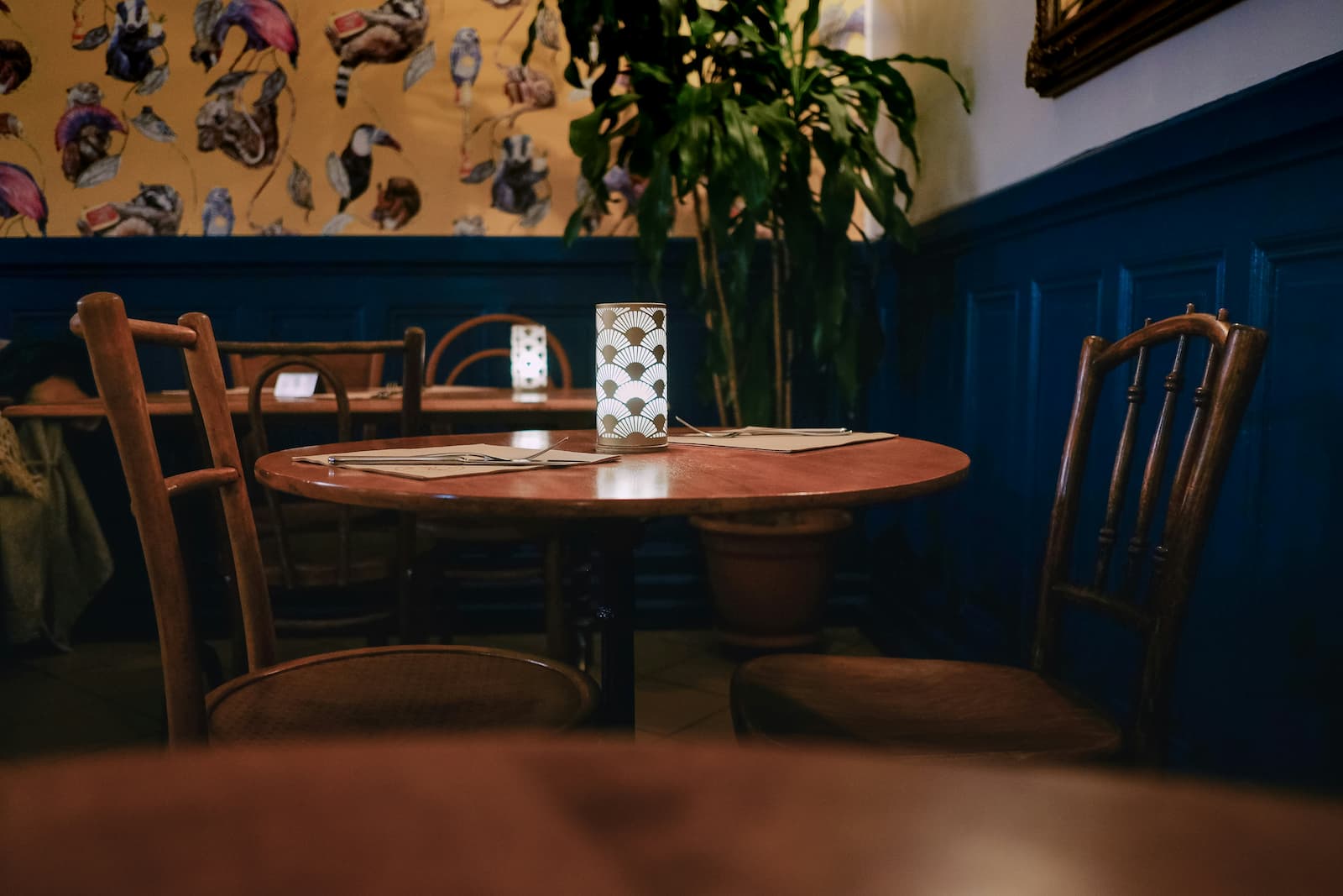 An empty restaurant table set with menus and a decorative lamp, surrounded by wooden chairs in a warmly lit dining room.