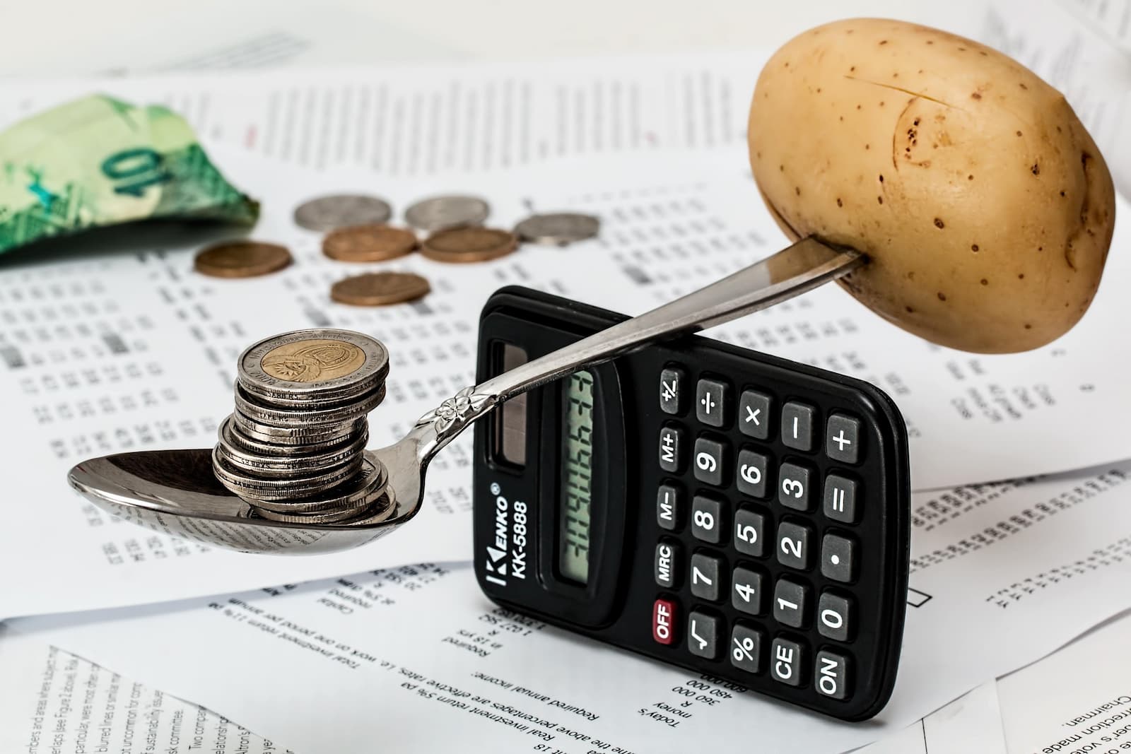 A potato balanced against stacked coins on spoons beside a calculator, banknotes, and financial documents, representing food costs and pricing.