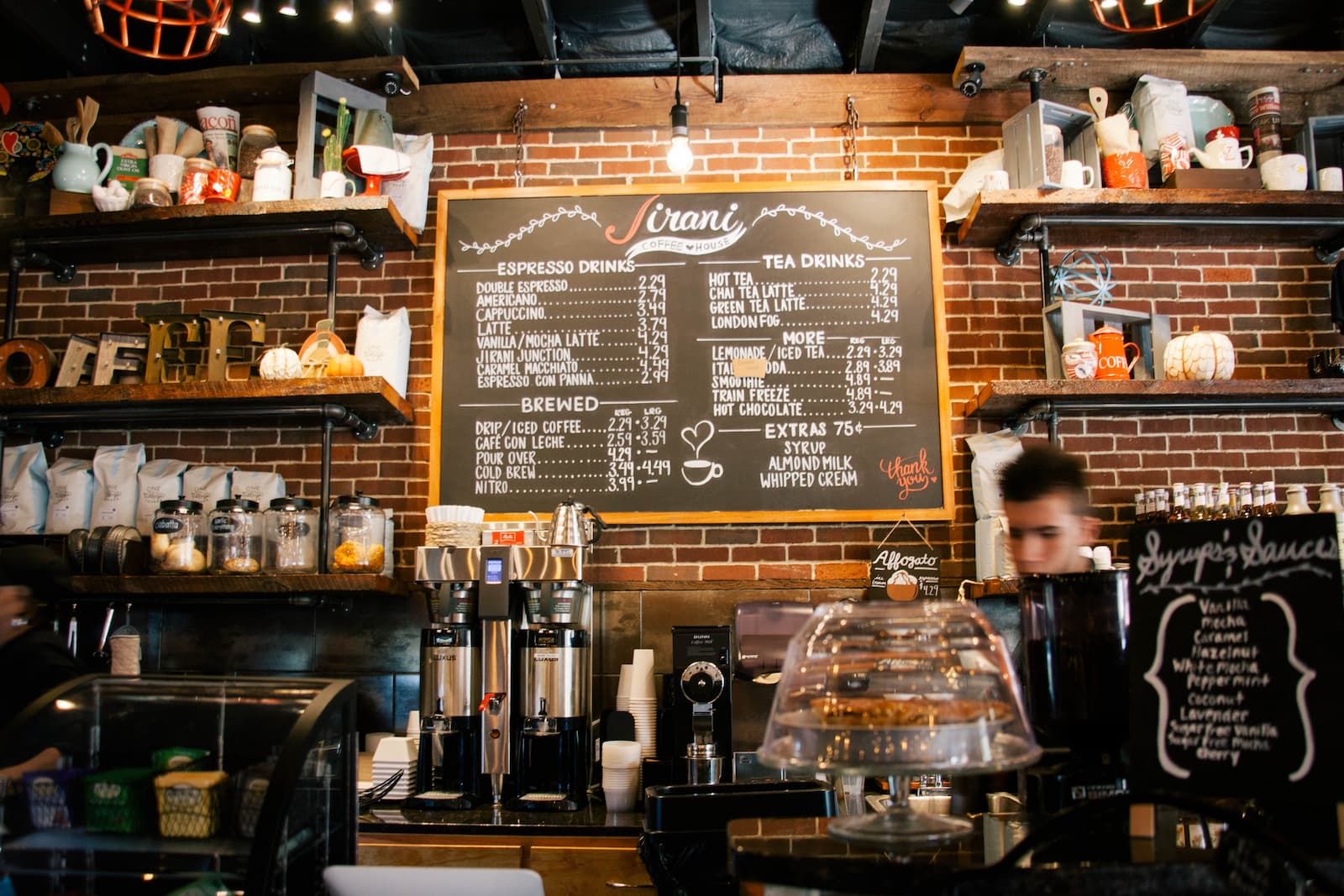 Interior of a coffee shop with a large chalkboard menu displaying espresso, tea, and other drinks above the service counter.