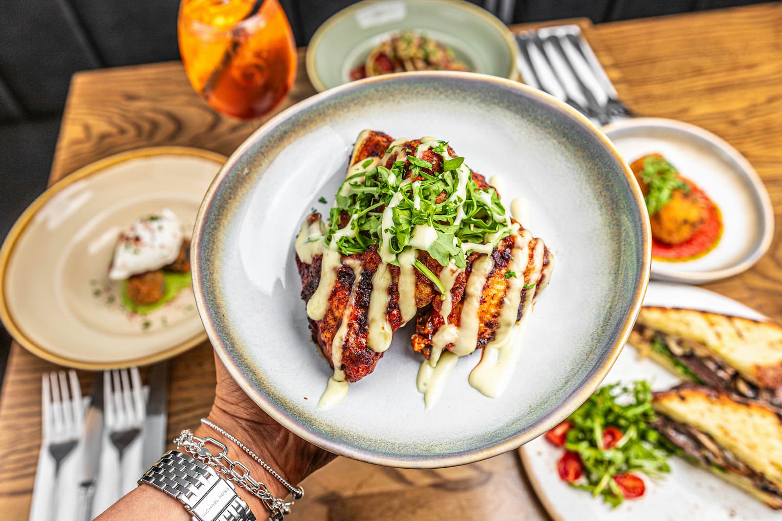 A hand holds a plated grilled dish topped with arugula and creamy sauce, surrounded by other restaurant plates.
